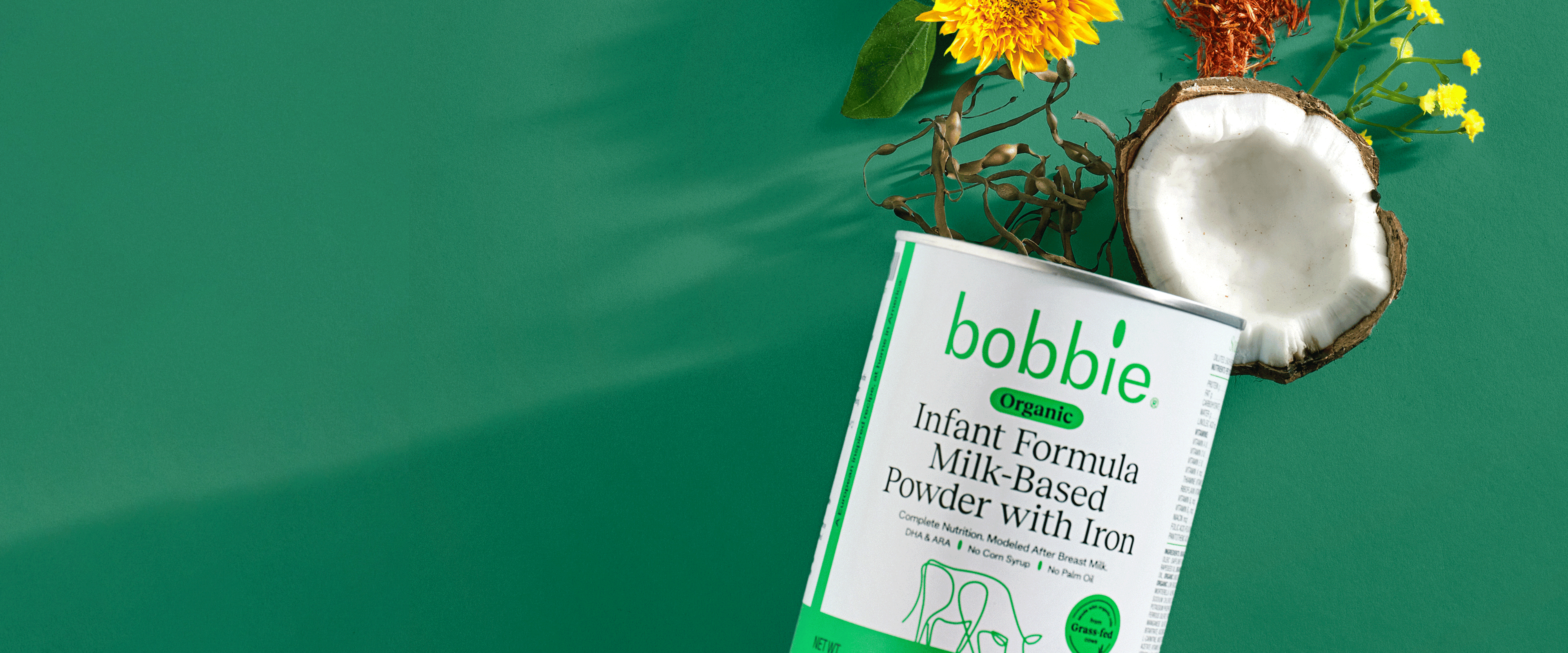 A can of organic infant formula with iron, surrounded by flowers and a coconut shell, set against a green background.